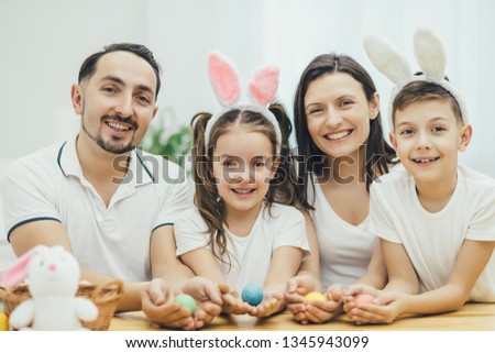 Inspired family in white t-shirts sitting together at the table, holding colored easter eggs in their hands, looking excitedly at the camera .