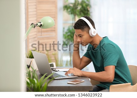 African-American teenage boy with headphones using laptop at table in room Royalty-Free Stock Photo #1346023922
