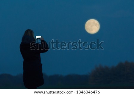 A woman seen as silhouette takes a picture of the full moon with her cell phone