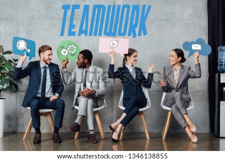 happy multiethnic businesspeople holding speech bubbles with teamwork icons while sitting in waiting hall