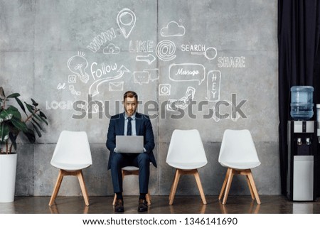 smiling businessman sitting on chair and using laptop in waiting hall with social media icons on wall