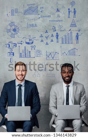smiling multiethnic businessmen looking at camera and using laptops in waiting hall with business development illustration on wall