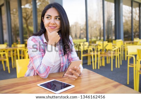Positive lady using tablet and smartphone in outdoor cafe. Pretty young woman sitting at table with chairs and building in background. Communication and technology concept. Front view.