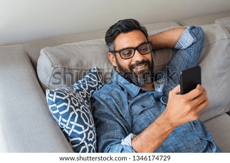 Happy smiling latin man using smartphone device while sitting on sofa at home. Mature indian man lying on couch reading messages on mobile phone. Hispanic guy with eyeglasses relaxing at home. Royalty-Free Stock Photo #1346174729