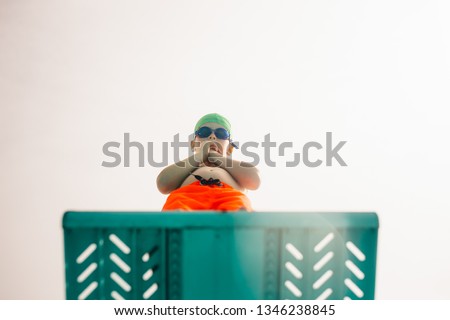 Low angle view of boy in swimming trunks, swim goggles and swimming cap standing on diving board against bright sky. Boy on diving platform at pool. Royalty-Free Stock Photo #1346238845
