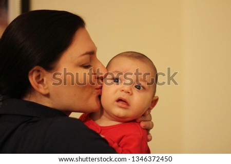 Young beautiful proud mother is kissing plump cheek of her little baby in red pajamas. Chubby kid looks confused and displeased a bit. 