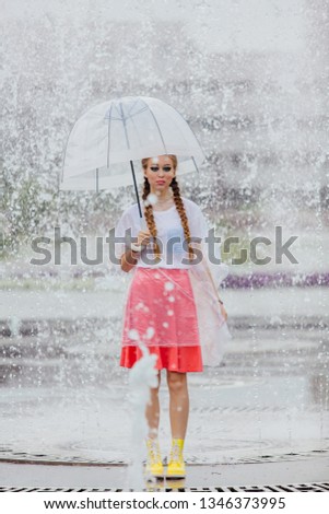 Young pretty girl with two braids in yellow boots and with transparent umbrella stands near fountain. Rainy day in city.