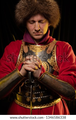 Medieval man in knight's armor and ancient red dress with furs in the yellow light on a dark background