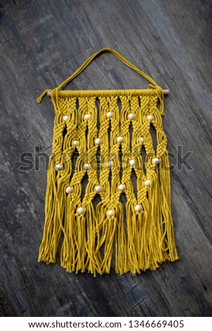 A yellow macrame laid onto a grey/brown wooden background. It's rectangular with yellow beads and the ends fraying.