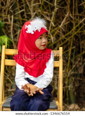 Muslim girls, islamic girls, wearing a red fabric head covering adorn with white fabric flowers red lip wear shirt white pants blue standing  photography on the chair prepare before to ramadan month.