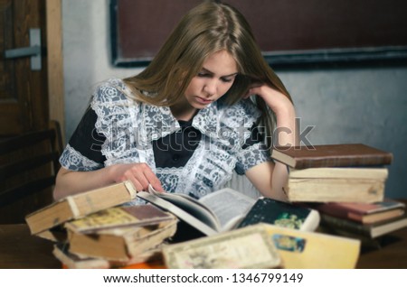 A young beautiful blond girl reads an old book. There are so many different old books on the table. She is dressed in school uniform and sits in classroom. Vintage photo