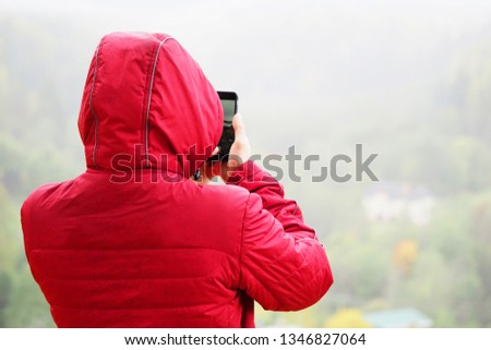 Man in a wet red jacket with a hood photographs on the phone the mountain landscape in the rain and fog. Outdoor shot in nature