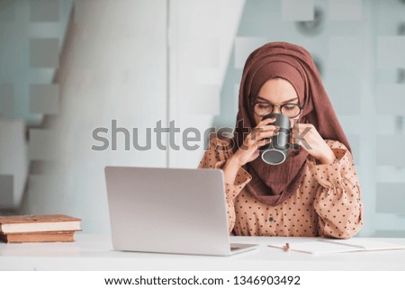 Young asian muslim woman sitting alone in a cafe working on her laptop and holding a coffee mug.