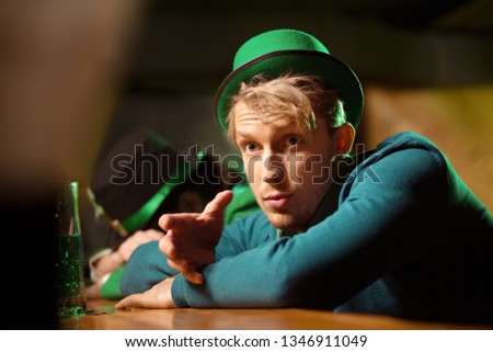Need more beer. Fair-haired young handsome man wearing a green looking surprised while sitting at the bar counter