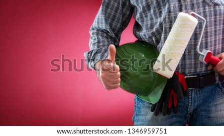 Caucasian house painter worker in jeans and shirt, with helmet and roller making ok sign with thumb up. Construction industry. Job security. Red background