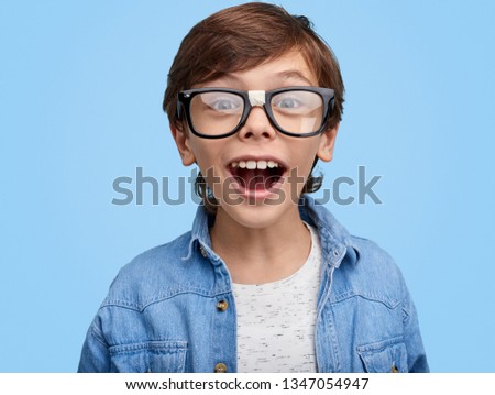 Happy nerdy boy in eyeglasses and denim jacket looking excitedly at camera on blue background . Closeup portrait of Excited school kid in broken glasses