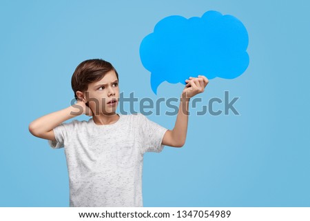 Casual little boy holding empty speech bubble and looking at it with confusion on blue background 