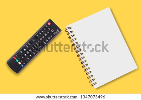 Black plastic remote control for tv near blank paper notepad with spiral binder lies on yellow table in home or office. Top view.Concept of communication or multimedia. Copy space for your text