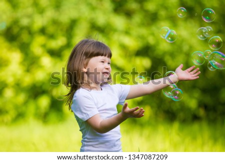 Beautiful little girl, has happy fun smiling face, pretty eyes, playing soap bubbles, dressed in white t-shirt, enjoying green forest. Child portrait. Summer activity rest. Kids fashion style. 