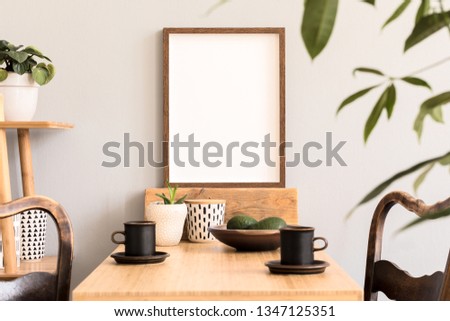 Stylish and sunny interior of kitchen space with wooden table with brown mock up photo frame, design chairs and bamboo shelf. Scandinavian room decor with kitchen accessories and beautiful plants.