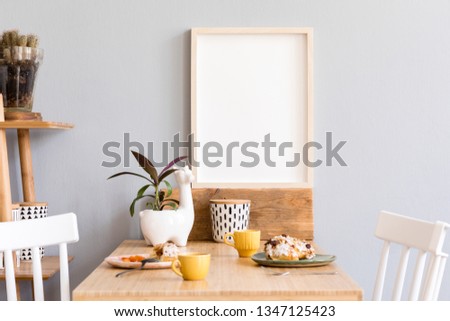 Stylish and sunny interior of kitchen space with small wooden table with mock up photo frame, design cups and tasty dessert. Scandinavian room decor with kitchen accessories, cacti and plants.