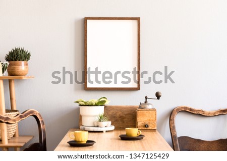 Retro and stylish interior of kitchen space with small wooden table with brown mock up photo frame, cups and vintage chairs. Scandinavian room decor with kitchen accessories and beautiful plants.