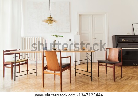 Eclectic and elegant dining room interior with design sharing table, chairs, gold pedant lamp, abstract paintings, piano and stylish accessories. Minimalistic decor. Brown wooden parquet. Real photo.