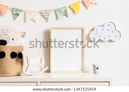Stylish and modern scandinavian childroom interior with mock up photo or poster frame on the white shelf. Toys, teddy bear in basket, rocking horse and hanging cotton colorful flags and cloud.