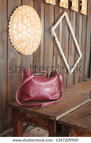 Woman's red leather bag is placed on a traditional wooden table and has a wooden wall as a backdrop. There are hats and picture frames on the wall.