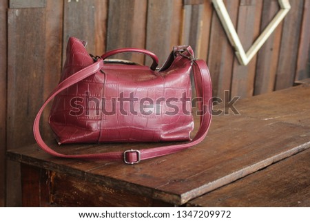 Woman's red leather bag is placed on a traditional wooden table and has a wooden wall as a backdrop. There are hats and picture frames on the wall.
