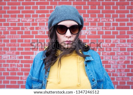 Young cute and pretty caucasian girl wearing yellow hoodie sweatshirt, denim jacket and a woolen hat, exterior photo shoot, posing in front of a red brick wall, spring or fall fashion concept 