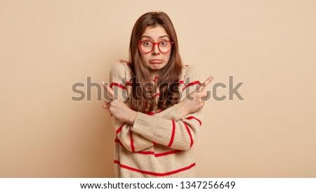 Horizontal shot of hesitant puzzled beautiful European woman crosses hands over chest, points both fore fingers at different corners, being unaware to choose something between, stands indoor