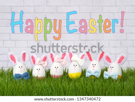 Egg craft bunnies, boys and girls lined up in a row on green grass next to a white brick wall with colorful Happy Easter text. Easter theme