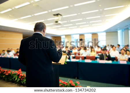Business man is making a speech in front of a big audience at a conference hall. Royalty-Free Stock Photo #134734202