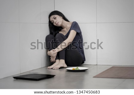Picture of unhealthy woman sitting with a plate of salad and weight scales in the bathroom