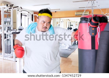 Picture of fat man wearing sportswear while punching a boxing sack in the gym center