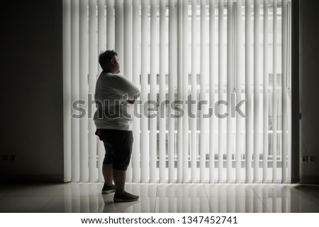 Picture of a lonely man looking out the window while standing in the dark room