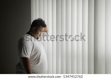 Picture of overweight man looks lonely while standing near the window in the dark room