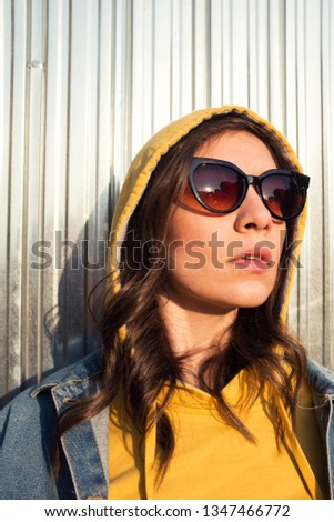 Young pretty caucasian girl wearing yellow hoodie sweater and denim jacket, exterior photo shoot, posing in front of a white metal wall, spring or fall fashion concept 
