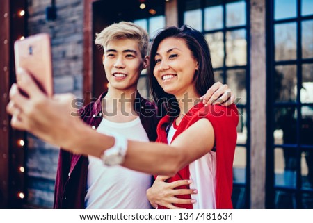 Cheerful chinese male and female bloggers holding smartphone and hugging while taking selfie pictures via front camera of device for social networks, concept of leisure time and relationship