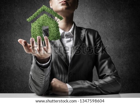 Businessman in suit presenting green plant in form of house symbol with gray wall on background.