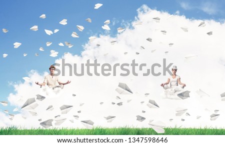 Young couple keeping eyes closed and looking concentrated while meditating on clouds among flying paper planes with bright and beautiful landscape on background.