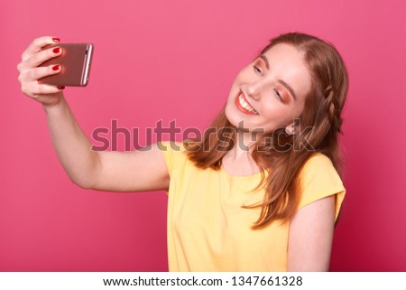 Close up portrait of young beautiful stylish women making selfie, using her own smartphone, smiles at camera, dressed casual yellow t shirt, has straight hair, wants new photo for social networks.