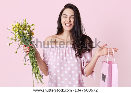 Close up portrait of cheerful female recieves present for birhday party, holds bag and flowers, looks happily, wears in dress, poses isolated over rosy background. Presents and celebration concept.