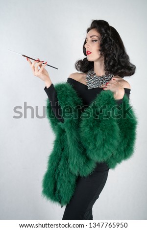 charming graceful pin up girl in a black evening dress and a green fluffy boa in her hands stands and smokes a cigarette with a mouthpiece on a white solid Studio background