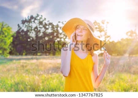 Happy smiling woman female in summer hat and lite yellow summer dress talking speaking by smartphone, mobile phone in green park outdoor. Summer, spring active outdoor leisure concept.