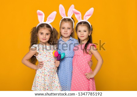 three little girls with Easter bunny ears holding colorful eggs on yellow background. Happy easter. cute children