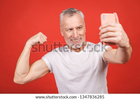 Portrait of happy senior bearded man showing biceps and taking selfie on smartphone, isolated against red background. Sport and healthy concept. 