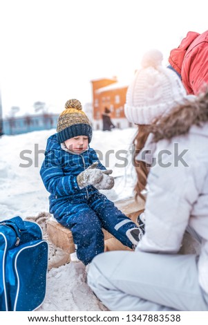 A small boy of 3-5 years old, sitting on a bench, in the winter in the city on the rink. Waiting for Mom to put on skates. On the cold day on weekends in January.