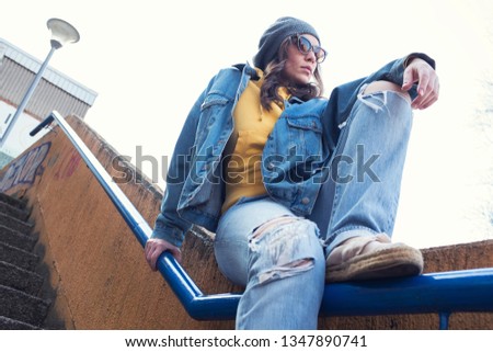 Young pretty caucasian girl wearing yellow hoodie sweater and denim jacket and jeans, exterior photo shoot, sitting on a metal staircase railing, low angle perspective, spring or fall fashion concept 